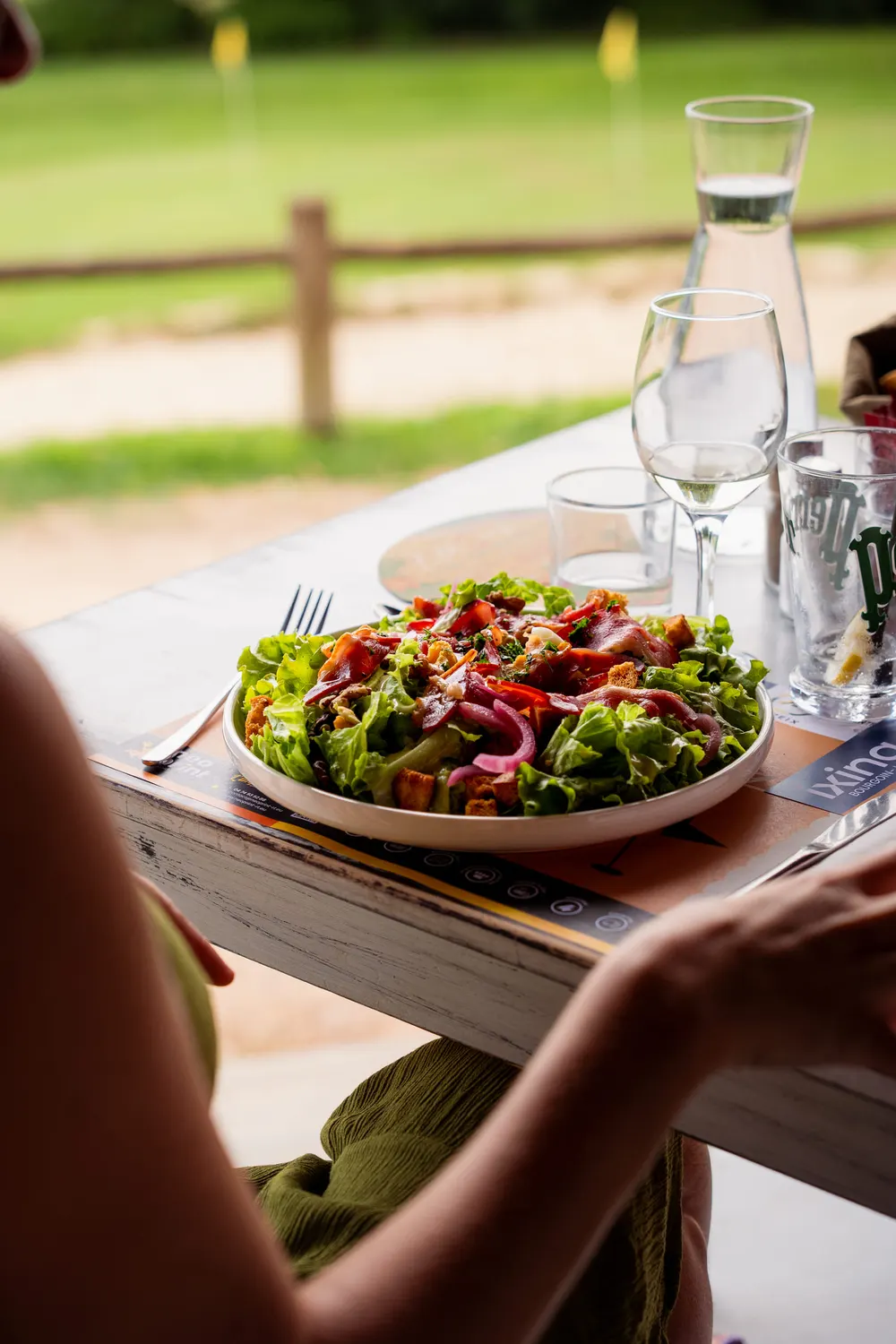 Grande salade servie en terrasse, carafe d’eau et vue sur le parcours